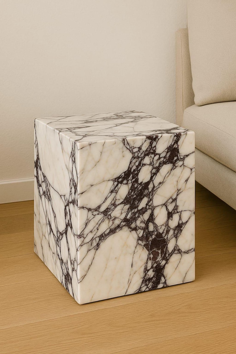 May include: A square marble side table with a white base and dark purple veining. The cube-shaped table is placed on a light wood floor, with a beige sofa in the background. The table is a modern piece of furniture.