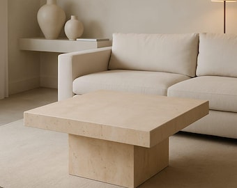 May include: A square, beige coffee table with a solid base and a flat top. The table is in front of a cream-colored sofa and a beige rug. The room also features a lamp and decorative vases.