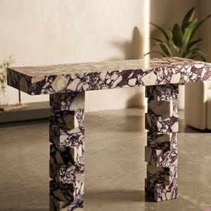 Puede incluir: Una mesa rectangular de mármol con un llamativo patrón morado y blanco. La parte superior de la mesa está sostenida por dos columnas, cada una construida con bloques rectangulares apilados. El diseño es moderno y la mesa probablemente se utiliza con fines decorativos.