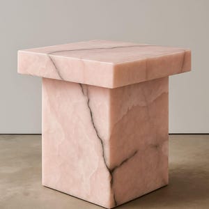 K&ouml;nnte beinhalten: Ein quadratischer, rosa Marmor-Beistelltisch mit rechteckiger Oberseite. Der Tisch hat eine massive, blockartige Basis und eine Oberseite mit leicht &uuml;berstehendem Rand. Dunkelgraue Aderung verleiht dem rosa Stein Details.