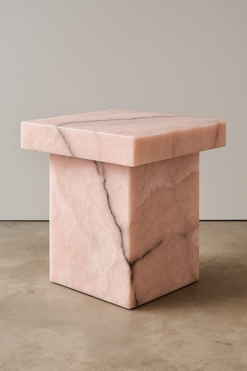 K&ouml;nnte beinhalten: Ein quadratischer, rosa Marmor-Beistelltisch mit rechteckiger Oberseite. Der Tisch hat eine massive, blockartige Basis und eine Oberseite mit leicht &uuml;berstehendem Rand. Dunkelgraue Aderung verleiht dem rosa Stein Details.