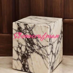 May include: A square marble side table with a white and dark brown veined pattern. The table is a cube shape and sits on a marble floor. The background is a dark wood panel.