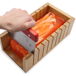 Può includere: Un tagliere per sapone in legno con una saponetta trasparente arancione e bianca all'interno. Una mano tiene un coltello di metallo con manico rosso e sta tagliando la saponetta.