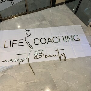 K&ouml;nnte beinhalten: Metallschild mit den Worten "LIFE COACHING meets Beauty" in einer Mischung aus Schriftarten. Eine Strichzeichnung einer Person ist integriert. Das Schild befindet sich auf einer wei&szlig;en Oberfl&auml;che, mit einem gefliesten Boden im Hintergrund.