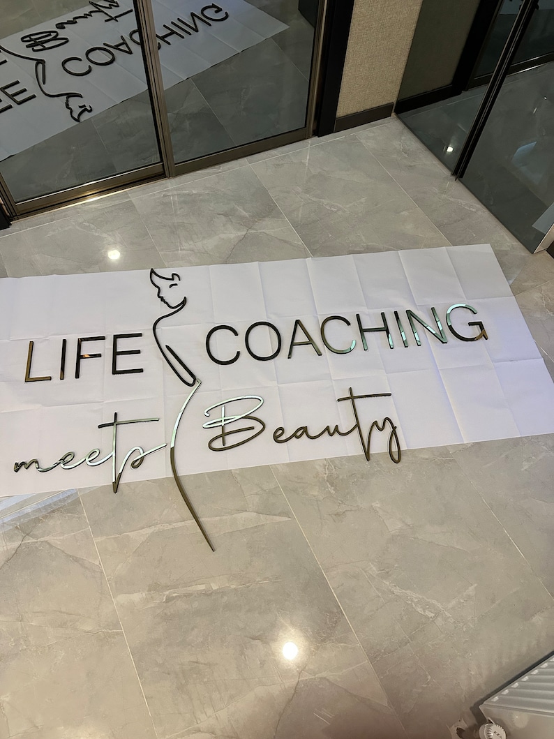 K&ouml;nnte beinhalten: Metallschild mit den Worten "LIFE COACHING meets Beauty" in einer Mischung aus Schriftarten. Eine Strichzeichnung einer Person ist integriert. Das Schild befindet sich auf einer wei&szlig;en Oberfl&auml;che, mit einem gefliesten Boden im Hintergrund.