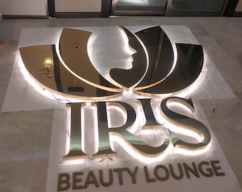 Könnte beinhalten: Ein großes, goldfarbenes und reflektierendes Schild mit einem stilisierten Lotusblüten-Symbol über dem Text IRIS und den Wörtern BEAUTY LOUNGE. Das Schild verfügt über eine integrierte Hintergrundbeleuchtung, die einen sanften Schein erzeugt.