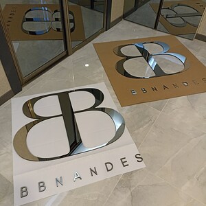K&ouml;nnte beinhalten: Zwei gro&szlig;e, reflektierende silberne Logos auf wei&szlig;en und braunen Hintergr&uuml;nden mit der Aufschrift "BBNANDES". Die Logos befinden sich auf einem gefliesten Boden, mit Spiegelt&uuml;ren im Hintergrund, die die Logos reflektieren. Die Buchstaben sind fett und ohne Serifen.