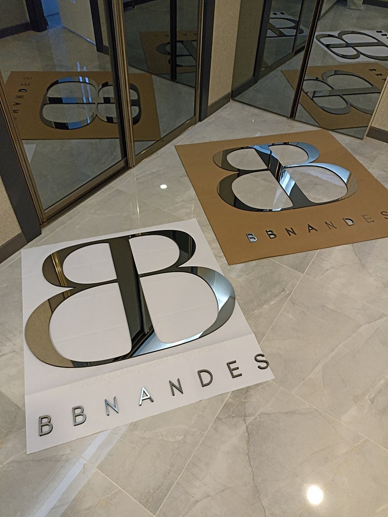 K&ouml;nnte beinhalten: Zwei gro&szlig;e, reflektierende silberne Logos auf wei&szlig;en und braunen Hintergr&uuml;nden mit der Aufschrift "BBNANDES". Die Logos befinden sich auf einem gefliesten Boden, mit Spiegelt&uuml;ren im Hintergrund, die die Logos reflektieren. Die Buchstaben sind fett und ohne Serifen.