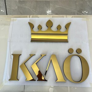 K&ouml;nnte beinhalten: Goldfarbene Krone und Buchstaben, die "KAO" buchstabieren, mit zwei Punkten &uuml;ber dem "O". Die Krone ist ein stilisiertes Design mit flacher Basis und mehreren Spitzen. Die Buchstaben sind in einer fetten, modernen Schriftart.