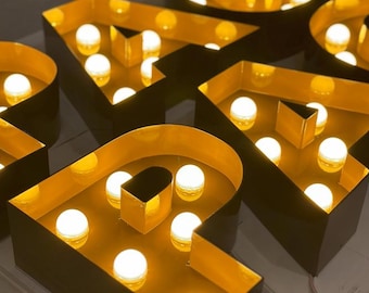 Könnte beinhalten: Nahaufnahme von beleuchteten Leuchtbuchstaben, die das Wort "PARADE" buchstabieren. Die Buchstaben sind goldfarben mit dunklem Rand, jede mit mehreren hellweißen Glühbirnen. Die Buchstaben sind auf einer hellen Oberfläche angeordnet.