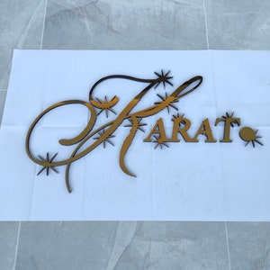K&ouml;nnte beinhalten: Ein goldfarbenes Schild mit dem Wort "FARAT" in Gro&szlig;buchstaben. Der Buchstabe "F" ist mit dekorativen Schn&ouml;rkeln und Sternen-Akzenten versehen. Das Schild ist wahrscheinlich f&uuml;r Unternehmen oder die Inneneinrichtung gedacht.