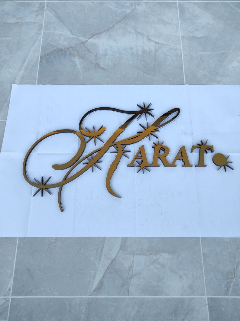K&ouml;nnte beinhalten: Ein goldfarbenes Schild mit dem Wort "FARAT" in Gro&szlig;buchstaben. Der Buchstabe "F" ist mit dekorativen Schn&ouml;rkeln und Sternen-Akzenten versehen. Das Schild ist wahrscheinlich f&uuml;r Unternehmen oder die Inneneinrichtung gedacht.