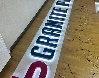 Könnte beinhalten: Ein langes, rechteckiges Schild mit den Worten "GRANITE PLUS" in großen, beleuchteten Buchstaben. Die Buchstaben sind schwarz mit weißem Leuchten, und das "G" und "A" sind rot. Elektrische Drähte sind mit dem Schild verbunden.