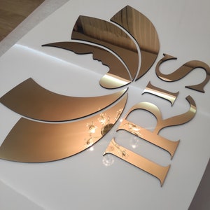 K&ouml;nnte beinhalten: Wanddekoration mit Spiegelgold-Design. Das Design umfasst eine stilisierte Bl&uuml;tenform und die Buchstaben "IRIS". Die Goldelemente sind vor einem wei&szlig;en Hintergrund platziert, was einen auff&auml;lligen Kontrast erzeugt. Die Oberfl&auml;che spiegelt das Licht.