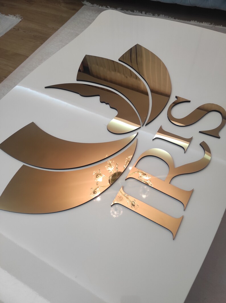 K&ouml;nnte beinhalten: Wanddekoration mit Spiegelgold-Design. Das Design umfasst eine stilisierte Bl&uuml;tenform und die Buchstaben "IRIS". Die Goldelemente sind vor einem wei&szlig;en Hintergrund platziert, was einen auff&auml;lligen Kontrast erzeugt. Die Oberfl&auml;che spiegelt das Licht.