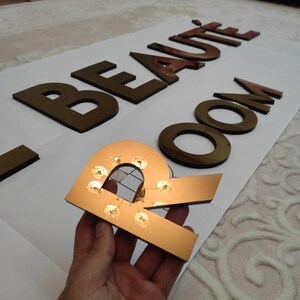 K&ouml;nnte beinhalten: Ein dekoratives Schild mit der Aufschrift "BEAUT&Eacute; ROOM" in goldfarbenen Buchstaben. Der Buchstabe "R" wird gehalten und verf&uuml;gt &uuml;ber kleine Lichter. Die Buchstaben sind auf einer wei&szlig;en Oberfl&auml;che platziert, was auf ein Beauty-Salon- oder Spa-Thema hindeutet.
