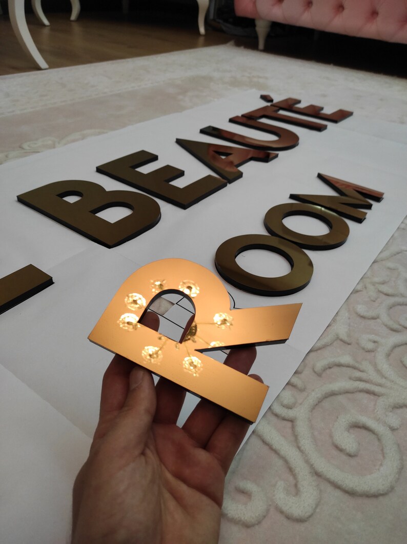 K&ouml;nnte beinhalten: Ein dekoratives Schild mit der Aufschrift "BEAUT&Eacute; ROOM" in goldfarbenen Buchstaben. Der Buchstabe "R" wird gehalten und verf&uuml;gt &uuml;ber kleine Lichter. Die Buchstaben sind auf einer wei&szlig;en Oberfl&auml;che platziert, was auf ein Beauty-Salon- oder Spa-Thema hindeutet.