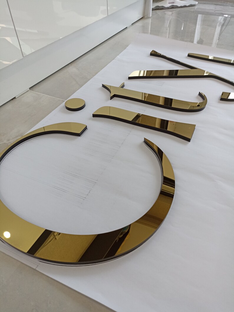 K&ouml;nnte beinhalten: Eine Sammlung goldfarbener, reflektierender Buchstaben und Formen, m&ouml;glicherweise f&uuml;r Schilder oder Dekoration. Die Teile sind auf einer wei&szlig;en Oberfl&auml;che angeordnet, mit einem kreisf&ouml;rmigen Element und verschiedenen Buchstabenformen. Die reflektierende Oberfl&auml;che erzeugt einen spiegelartigen Effekt.