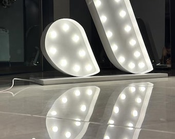 Könnte beinhalten: Weiße Leuchtbuchstaben im Marquee-Stil, die den Buchstaben "A" bilden. Die Lichter sind eingeschaltet und erzeugen einen warmen Schein. Die Buchstaben stehen auf einer reflektierenden Oberfläche, wodurch ein Spiegeleffekt entsteht. Das Design ist modern und dekorativ.