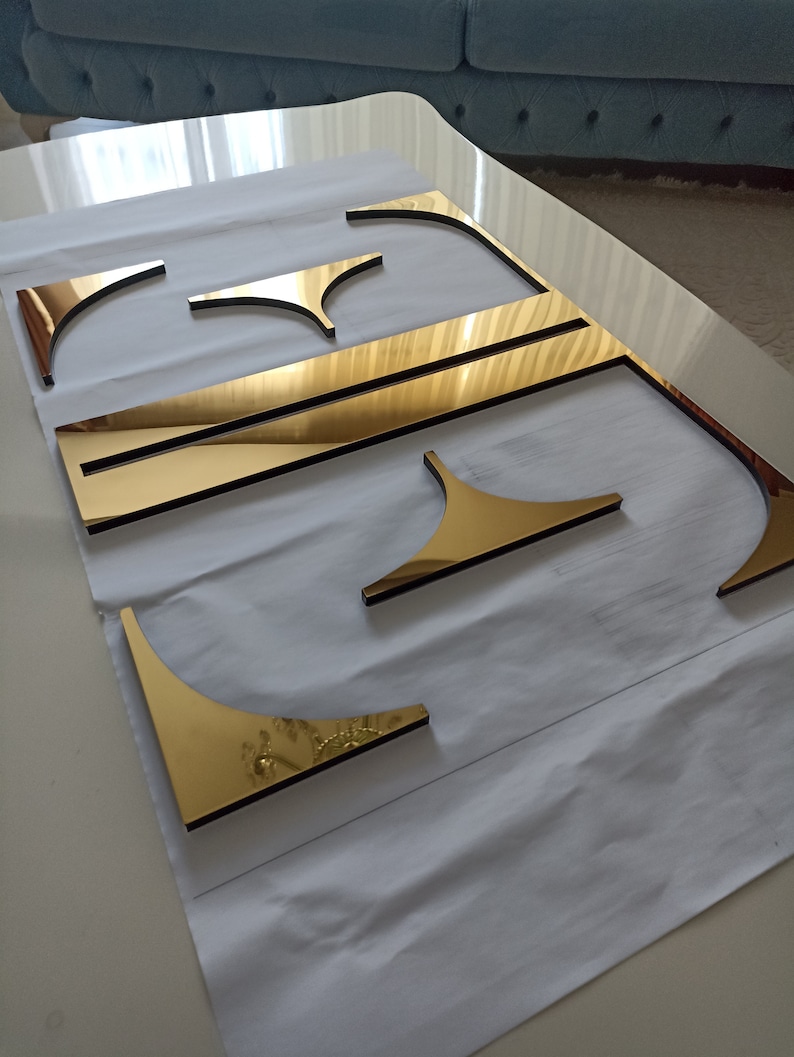 K&ouml;nnte beinhalten: Goldfarbene Metallbuchstaben "E"-Ausschnitte mit reflektierender Oberfl&auml;che, angeordnet auf einer wei&szlig;en Oberfl&auml;che. Der Buchstabe besteht aus mehreren Teilen, darunter gebogene und gerade Segmente sowie dekorative Elemente.