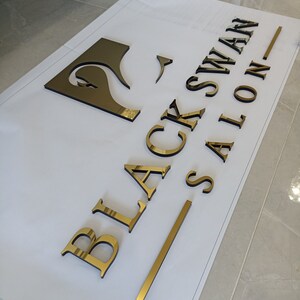 K&ouml;nnte beinhalten: Goldfarbene, dreidimensionale Buchstaben und ein Schwan-Logo, die "BLACK SWAN SALON" auf einer wei&szlig;en Oberfl&auml;che buchstabieren. Die Buchstaben sind reflektierend und scheinen aus Metall zu sein. Das Schwan-Logo ist eine stilisierte Silhouette.