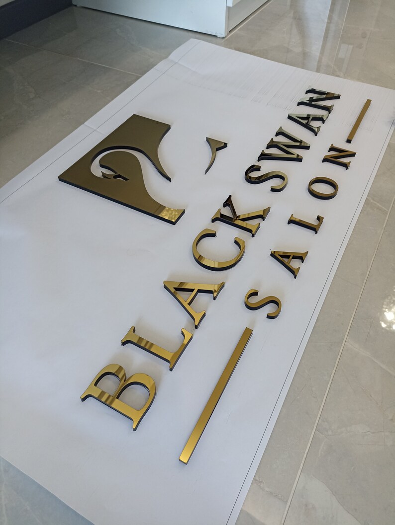 K&ouml;nnte beinhalten: Goldfarbene, dreidimensionale Buchstaben und ein Schwan-Logo, die "BLACK SWAN SALON" auf einer wei&szlig;en Oberfl&auml;che buchstabieren. Die Buchstaben sind reflektierend und scheinen aus Metall zu sein. Das Schwan-Logo ist eine stilisierte Silhouette.