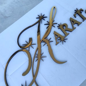 K&ouml;nnte beinhalten: Goldfarbenes, lasergeschnittenes Holzschild mit dem Wort "KRAT" und einem stilisierten Buchstaben "K". Der Buchstabe "K" ist mit Sternen-Akzenten verziert. Das Schild dient wahrscheinlich dekorativen Zwecken.