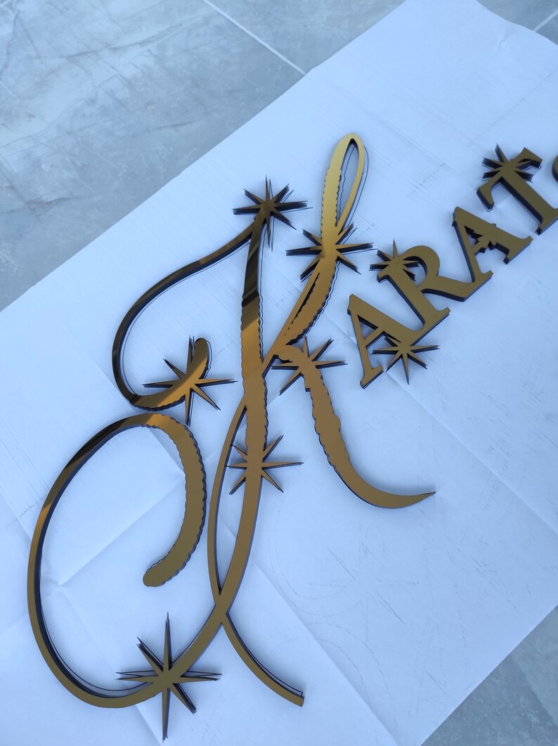 K&ouml;nnte beinhalten: Goldfarbenes, lasergeschnittenes Holzschild mit dem Wort "KRAT" und einem stilisierten Buchstaben "K". Der Buchstabe "K" ist mit Sternen-Akzenten verziert. Das Schild dient wahrscheinlich dekorativen Zwecken.