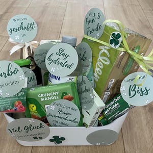 以下が含まれることがあります： スナック菓子、飲み物、パーソナルケア製品など、さまざまなアイテムが入った白いギフトバスケット。バスケットには、水筒、Kleenexの箱、Crunchy Mandelチョコレートバーが入っています。テキストとクローバーのデザインが施された丸いタグがいくつか付いています。