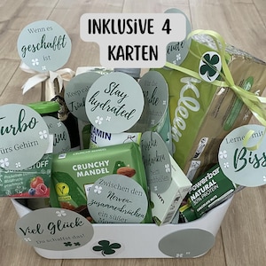 Op de afbeelding: Een cadeaupakket gevuld met diverse items, waaronder snacks, drankjes en kaarten. De mand bevat een verscheidenheid aan producten, waaronder een groene reep Crunchy Mandel, een Powerbar en andere verpakte goederen. De mand heeft verschillende ronde kaarten met tekst.