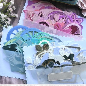 May include: A collection of paper die cuts in various pastel colors, including pink, blue, and green, arranged in white scalloped-edge envelopes. The die cuts feature floral, geometric, and decorative shapes, ideal for scrapbooking or card making.