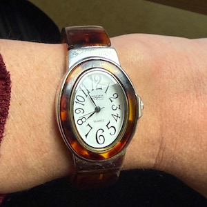 Könnte beinhalten: Ovale Quarz-Uhr mit silberfarbener Lünette und einem Rahmen mit Schildpattmuster. Das weiße Zifferblatt hat schwarze Ziffern und Zeiger. Das Armband ist braun.