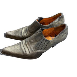 以下が含まれることがあります： メタリックゴールドのスリッポンシューズのペア。ポインテッドトゥ。靴は、質感のある、使い古された外観と低いヒールを持っています。内側はオレンジ色です。靴は視聴者の方を向いています。