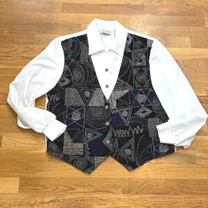 May include: A white long-sleeved blouse with a patterned vest. The vest features a geometric design in black, navy, gray, and teal. The blouse has a collared neckline and gold buttons. The vest is sleeveless and has a V-neck.