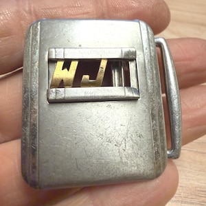 Può includere: Una fibbia per cintura rettangolare in metallo color argento con una finestra centrale ritagliata orizzontale. All'interno di questa finestra sono posizionate due lettere in metallo color oro, W e J. La superficie presenta segni di usura e graffi.
