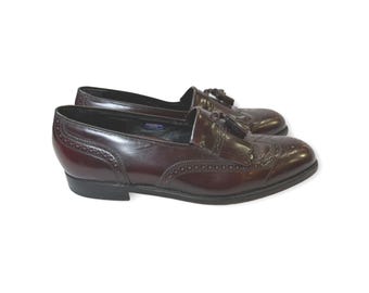 Pode incluir: Um par de mocassins de couro bordô com perfurações decorativas e detalhes de borla. Os sapatos têm um salto baixo e um acabamento polido, adequados para trajes formais ou de negócios.