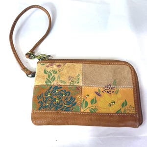 May include: A patchwork wristlet wallet with a brown leather wrist strap. The wallet features a variety of floral patterns in shades of yellow, green, and brown. Gold-colored studs accent the floral designs. The wallet has a brown zipper.