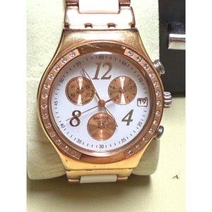 May include: A rose gold-toned Swatch watch with a white face, featuring gold-toned subdials and a date display. The bezel is adorned with small, clear crystals. The watch has a white leather strap and a rose gold-toned metal band.