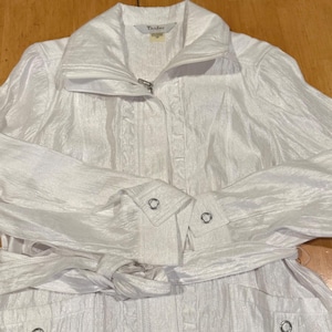 May include: A white jacket with a zip-up front, a collar, and long sleeves. The jacket has a tie belt and buttoned cuffs. The brand name "Tailday" is visible on the label. The jacket is made of a shiny, textured fabric.