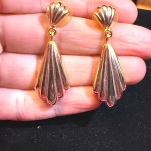 May include: A pair of gold-coloured earrings. Each earring has a scalloped top and a fan-shaped drop. The earrings are designed with a textured, ridged pattern, and are secured with a post backing. The earrings are displayed against a neutral background.