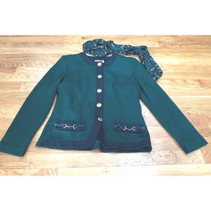 May include: A dark green long-sleeved jacket with a round neckline, a dark navy blue front placket featuring five light-colored buttons, and two front pockets adorned with silver-toned horse bit hardware, accompanied by a matching green scarf.