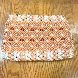 May include: A rectangular macrame clutch in shades of orange and white. The clutch features a geometric diamond pattern with a zipper closure. The bag is made of woven cord and has a textured appearance.