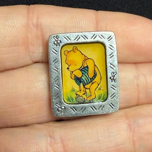 May include: A rectangular silver-toned metal pin featuring a central illustration of a yellow bear holding a blue striped honey pot. The frame has a textured border with etched diagonal lines and small bee motifs on the left and right sides.