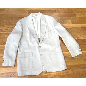 Könnte beinhalten: Ein hellbeiger Blazer mit steigendem Revers, zwei goldfarbenen Knöpfen als Verschluss und zwei großen aufgesetzten Taschen. Die Ärmel sind mit drei kleineren goldfarbenen Knöpfen an den Manschetten versehen und das Innere ist weiß gefüttert.