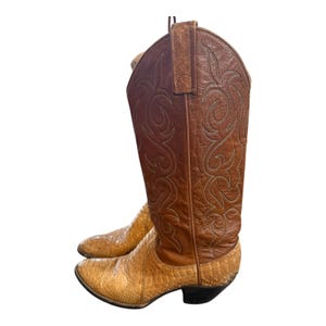 May include: A pair of cowboy boots. The boots are a light brown color with a snakeskin pattern on the foot and a darker brown leather upper. The upper features decorative stitching. The boots have a pointed toe and a black heel.