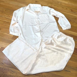 Könnte beinhalten: Ein zweiteiliger Schlafanzug in blassem Cremeton mit satinartiger Oberfläche. Das langärmelige Hemd mit Knopfleiste hat einen Kragen und eine einzelne Brusttasche mit gestepptem Rautenmuster. Die passende lange Hose verfügt über einen Gummibund.