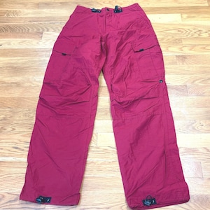 May include: Deep red cargo pants featuring a button and zipper fly closure, two large side cargo pockets with flaps, and adjustable drawstring toggles at the waist and the bottom of each pant leg. The pants have a relaxed fit and straight legs.