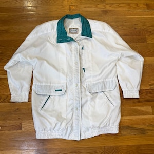 May include: White jacket with a teal green collar, pocket accents, and a small label. The jacket has a button-up front, two front pockets, and long sleeves. The jacket is made of a lightweight material.