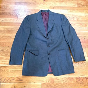 Könnte beinhalten: Ein anthrazitfarbener Blazer mit einer Drei-Knopf-Vorderseite, fallendem Revers und langen Ärmeln mit geknöpften Manschetten. Das Innere ist mit einem tief burgunderroten Stoff gefüttert, und die Außenseite verfügt über zwei Pattentaschen.