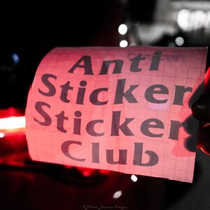 Puede incluir: Una pegatina rosa con el texto "Anti Sticker Club" en letras negras. La pegatina está sobre un fondo cuadriculado.